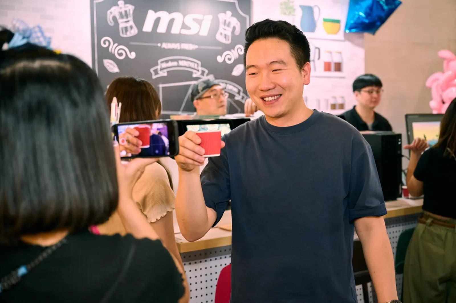 2023 女生日常節現場的 msi 品牌攤位，一位參加者舉起品牌飲品杯，另一位正用手機拍攝